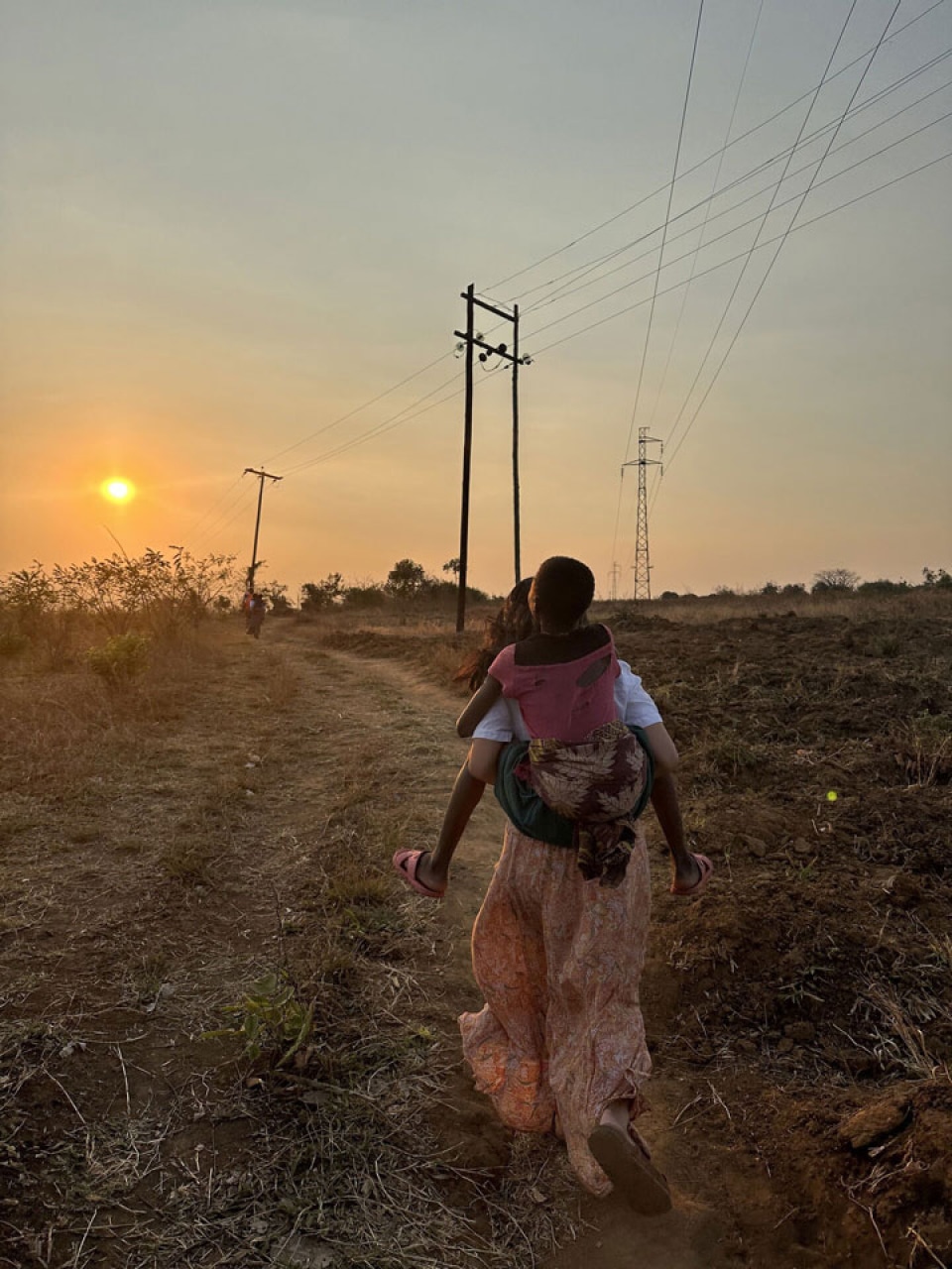 a person carrying a child on her back walking on a dirt path